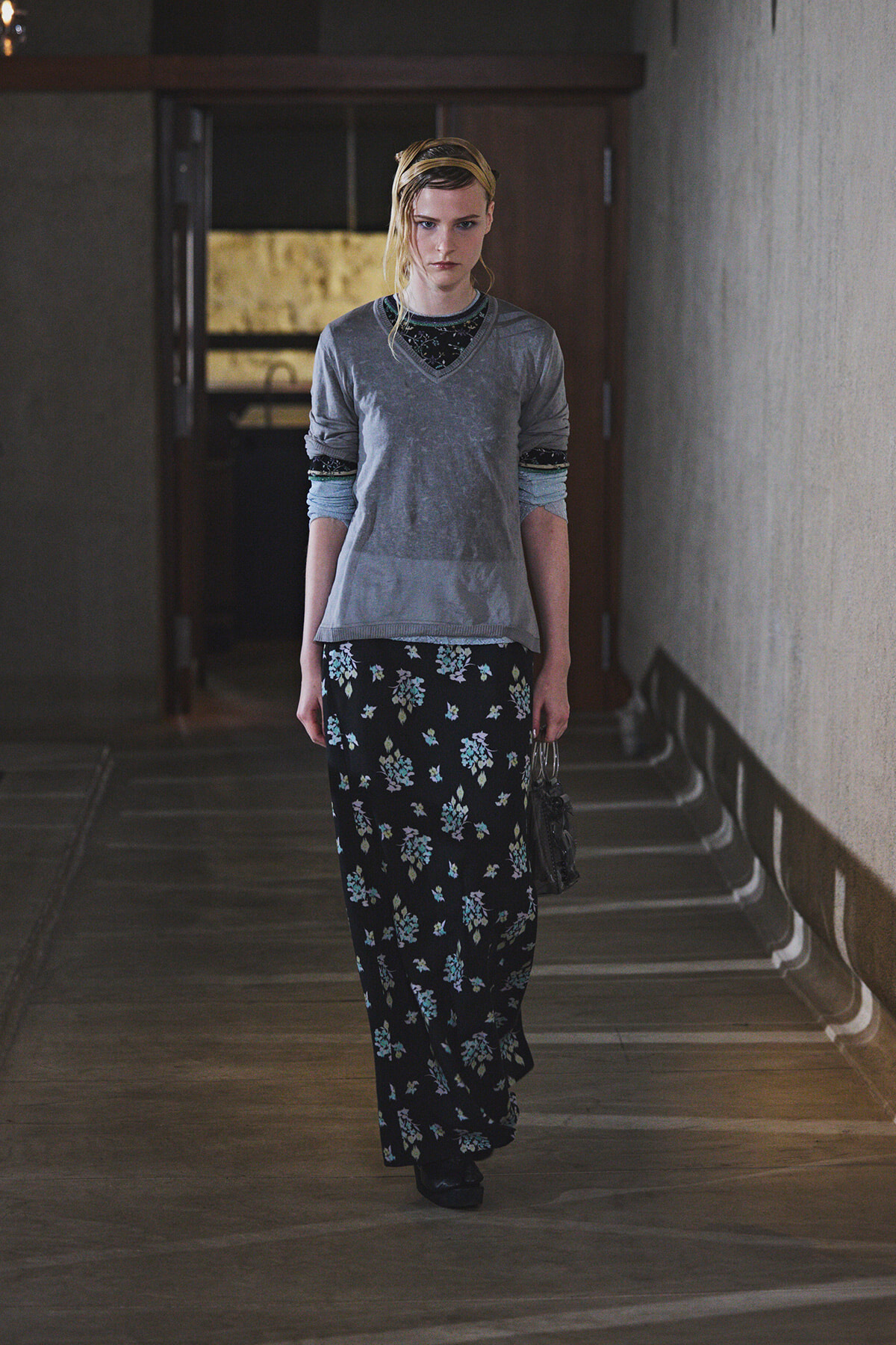 Woman walking down a dim hallway in a gray V-neck top over a black floral maxi skirt, carrying a small bag.