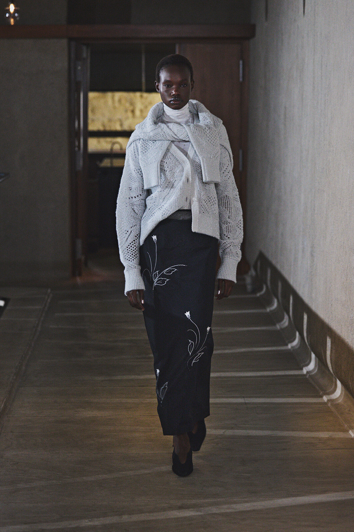 Person walking down a dim hallway wearing a chunky white knit cardigan draped over shoulders and a black floral-embroidered skirt.