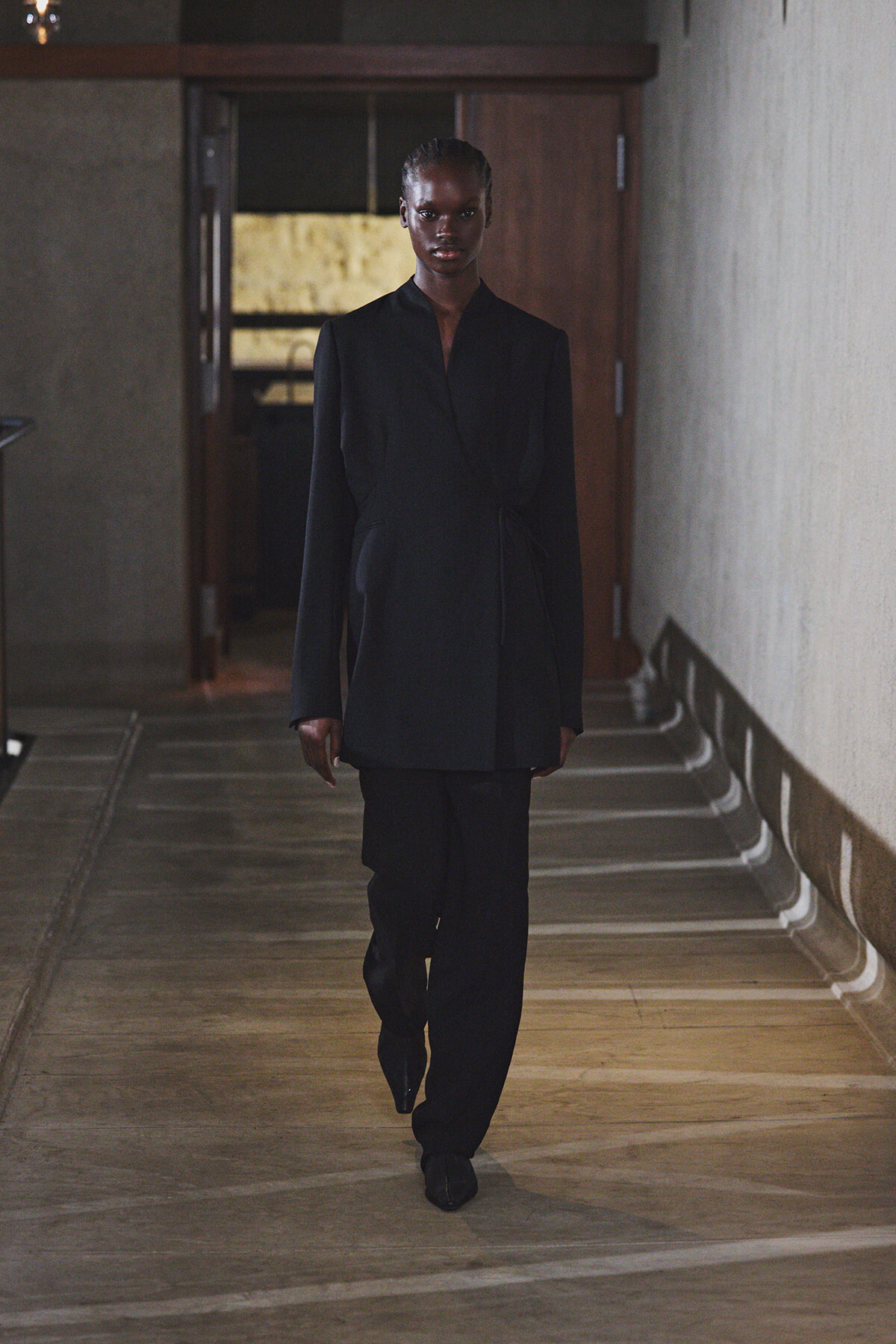 A person in a black tailored wrap blazer and matching pants walks down a dimly lit hallway.