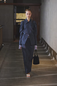 Young woman in a dark blue tailored suit with embroidered details walks down a dim hallway, carrying a small black handbag.