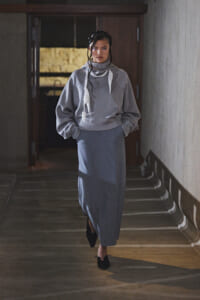 Woman in a gray oversized hoodie and long blue skirt walking down a dim hallway, hands in pockets.