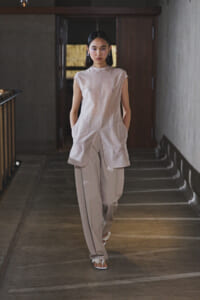 Model walking down a dim hallway in a loose beige sleeveless tunic and wide-leg pants, white flower-adorned sandals.