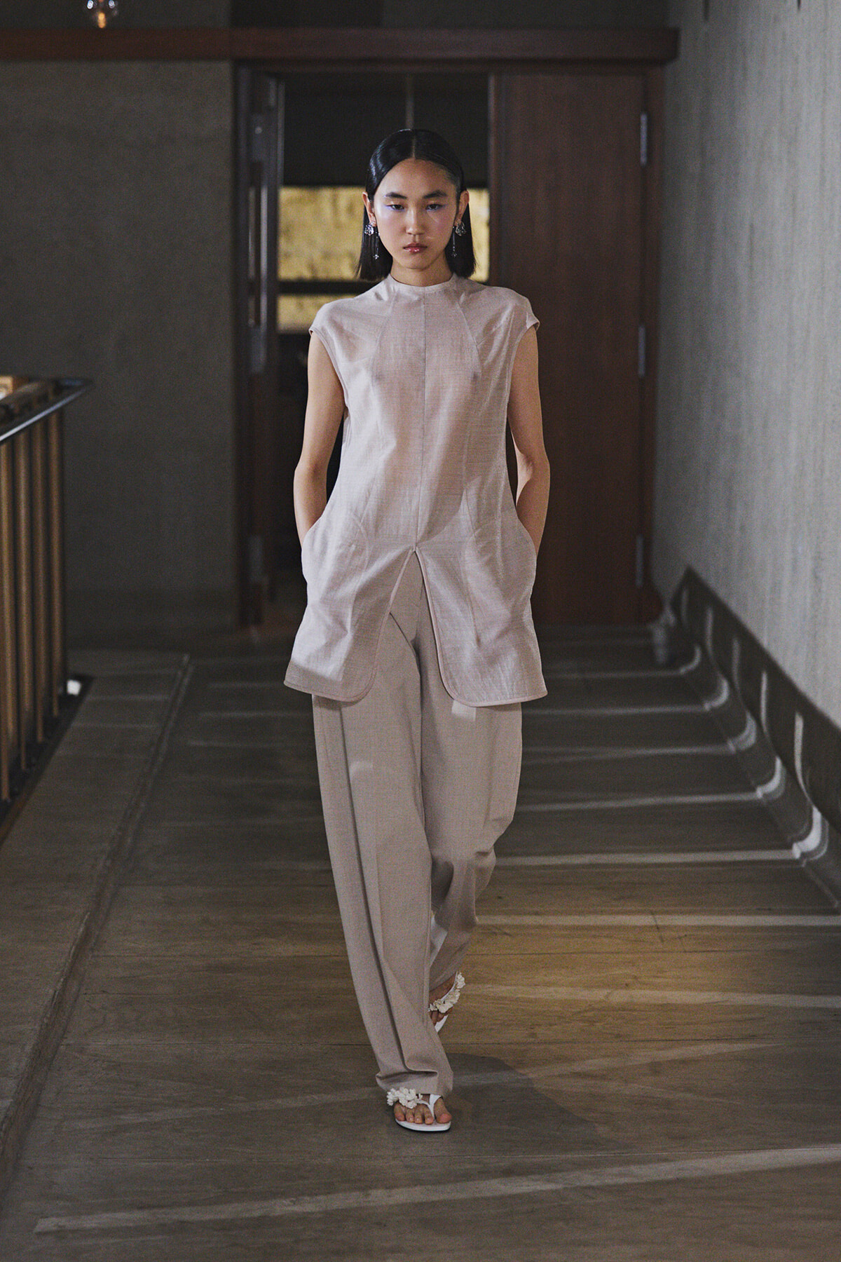 Model walking down a dim hallway in a loose beige sleeveless tunic and wide-leg pants, white flower-adorned sandals.