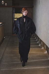 Model wearing an oversized black bomber jacket and long black skirt walking toward the camera in a dim hallway.