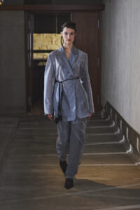 Model walks down a dim corridor in a light gray wrap blazer and matching pants, with a black belt sash.