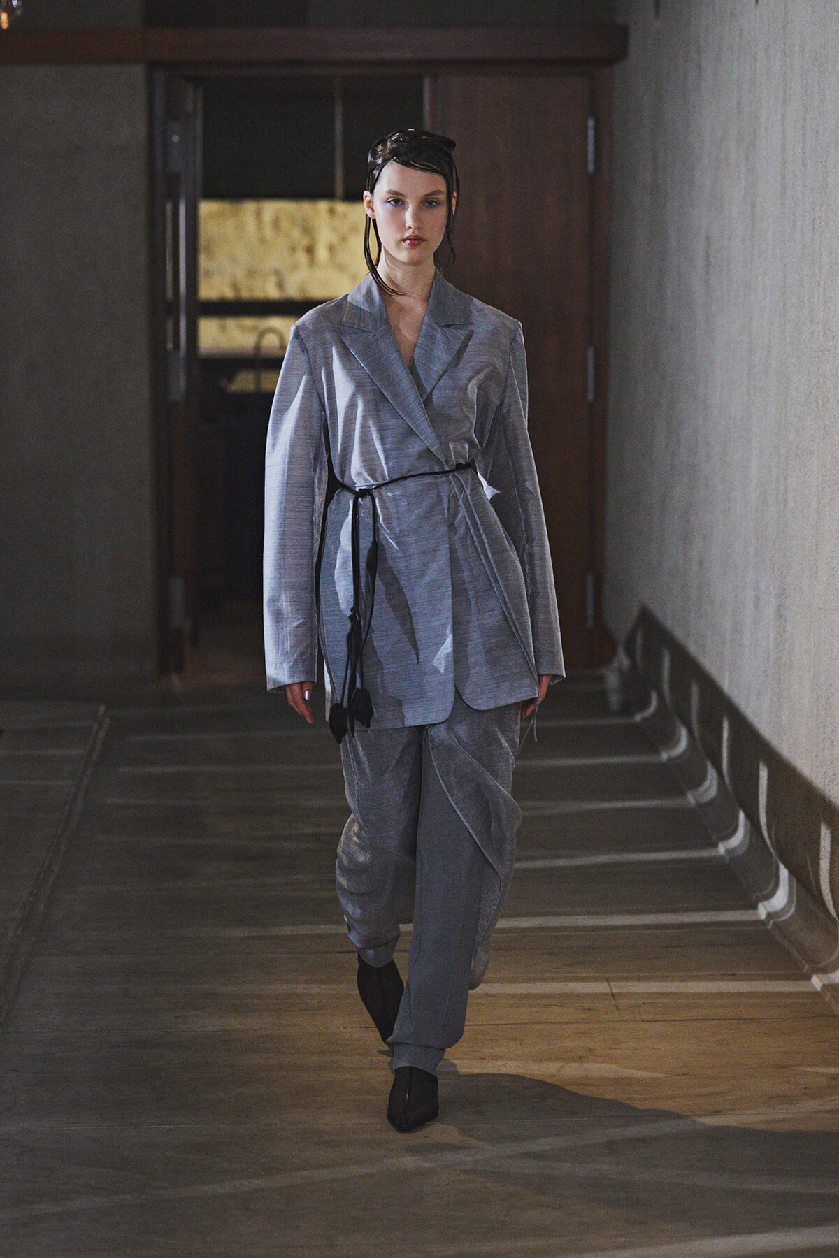Model walks down a dim corridor in a light gray wrap blazer and matching pants, with a black belt sash.