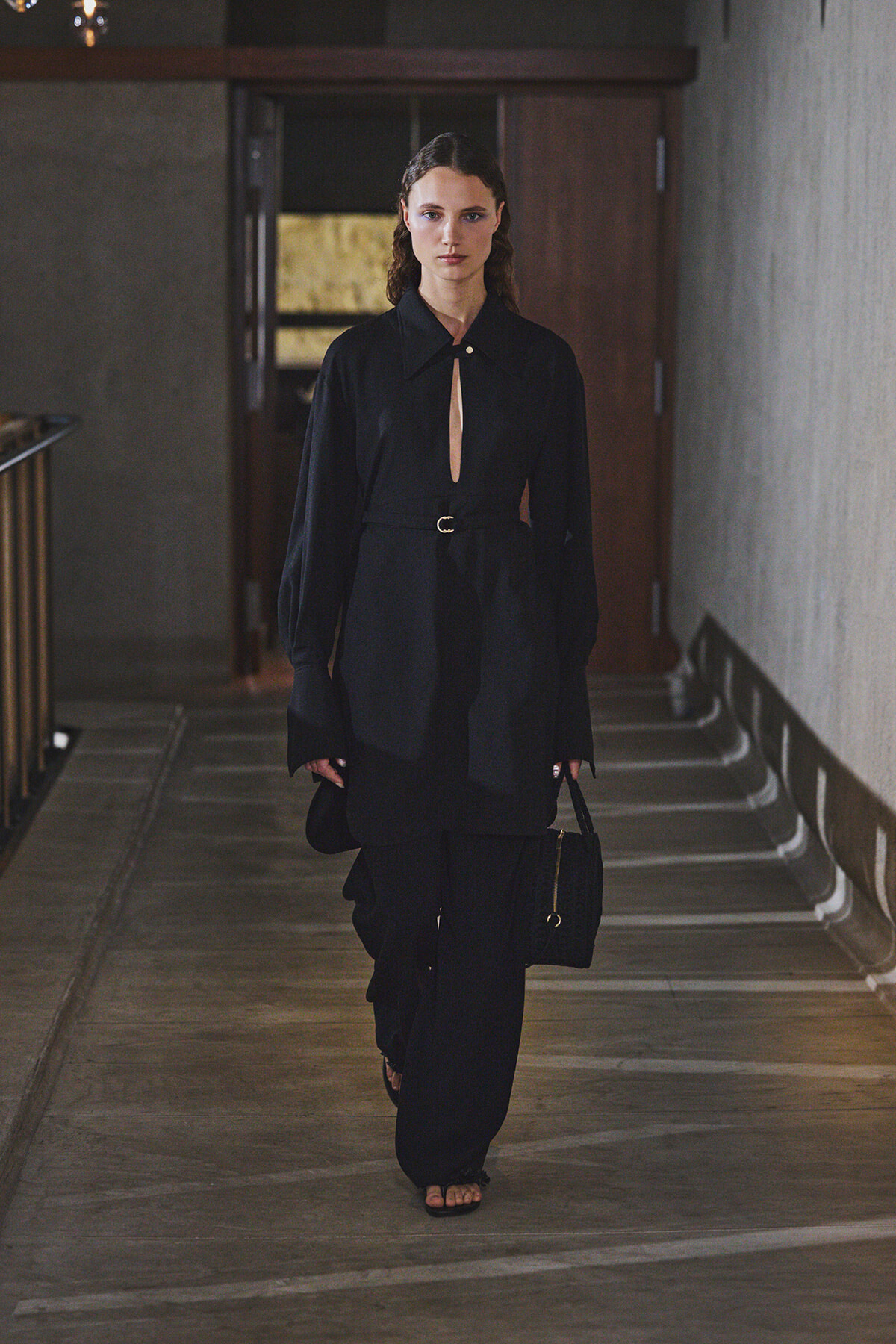 Woman in a black belted coat and wide-leg pants walks down a dim hallway, carrying a black bag.