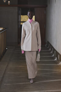 Model walks down a dim hallway wearing a beige pantsuit with a pink turtleneck and carries a small handbag.