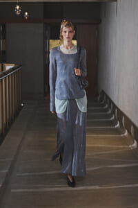 Model walking down a dim indoor hallway in a blue-gray star-patterned outfit, carrying a black quilted bag with a chain strap.