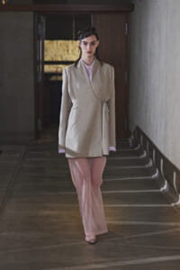 Woman model walking toward the camera in a light beige wrap blazer tied at the side and pale pink wide-leg pants, in a dim hallway.