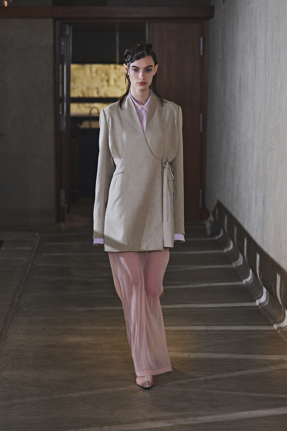 Woman model walking toward the camera in a light beige wrap blazer tied at the side and pale pink wide-leg pants, in a dim hallway.