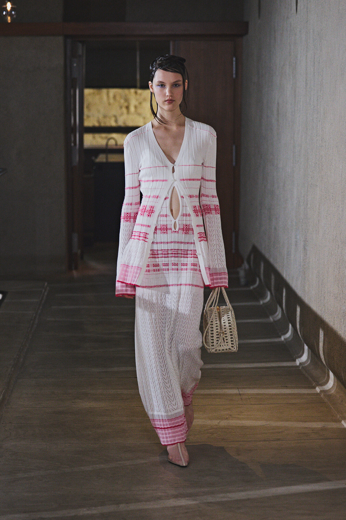 Model walking in a white and pink knit cardigan and matching pants, carrying a woven handbag down a runway.