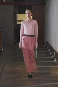 Model walking down a dim hallway in a pale pink V-neck sweater and a matching pink skirt with a black waistband, carrying a small beaded handbag.
