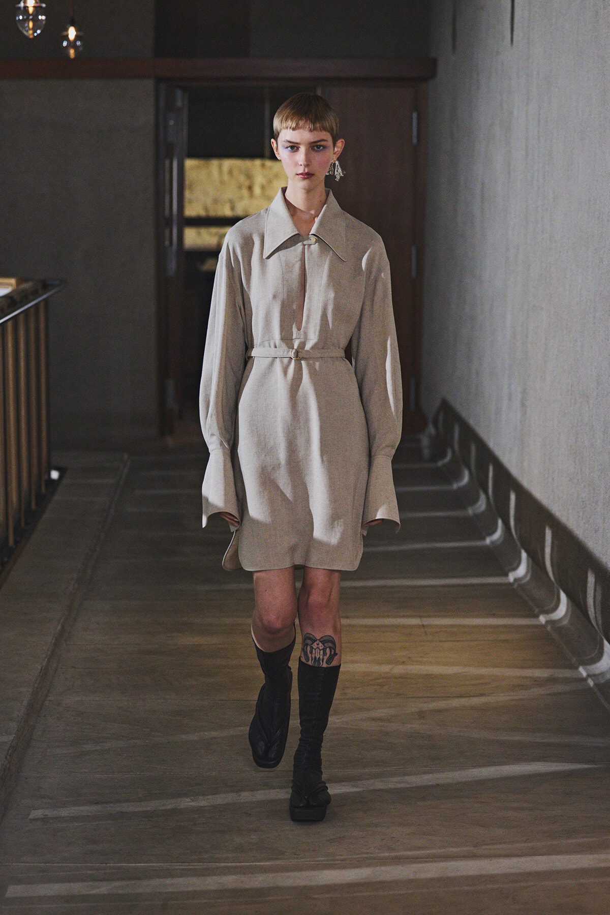 Model walks down a dim hallway in a beige shirt-dress with a belt, black knee-high boots, and a visible leg tattoo.
