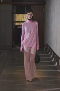 Model in a light pink, long-sleeve, high-neck dress walking down a dim hallway with a small black handbag.