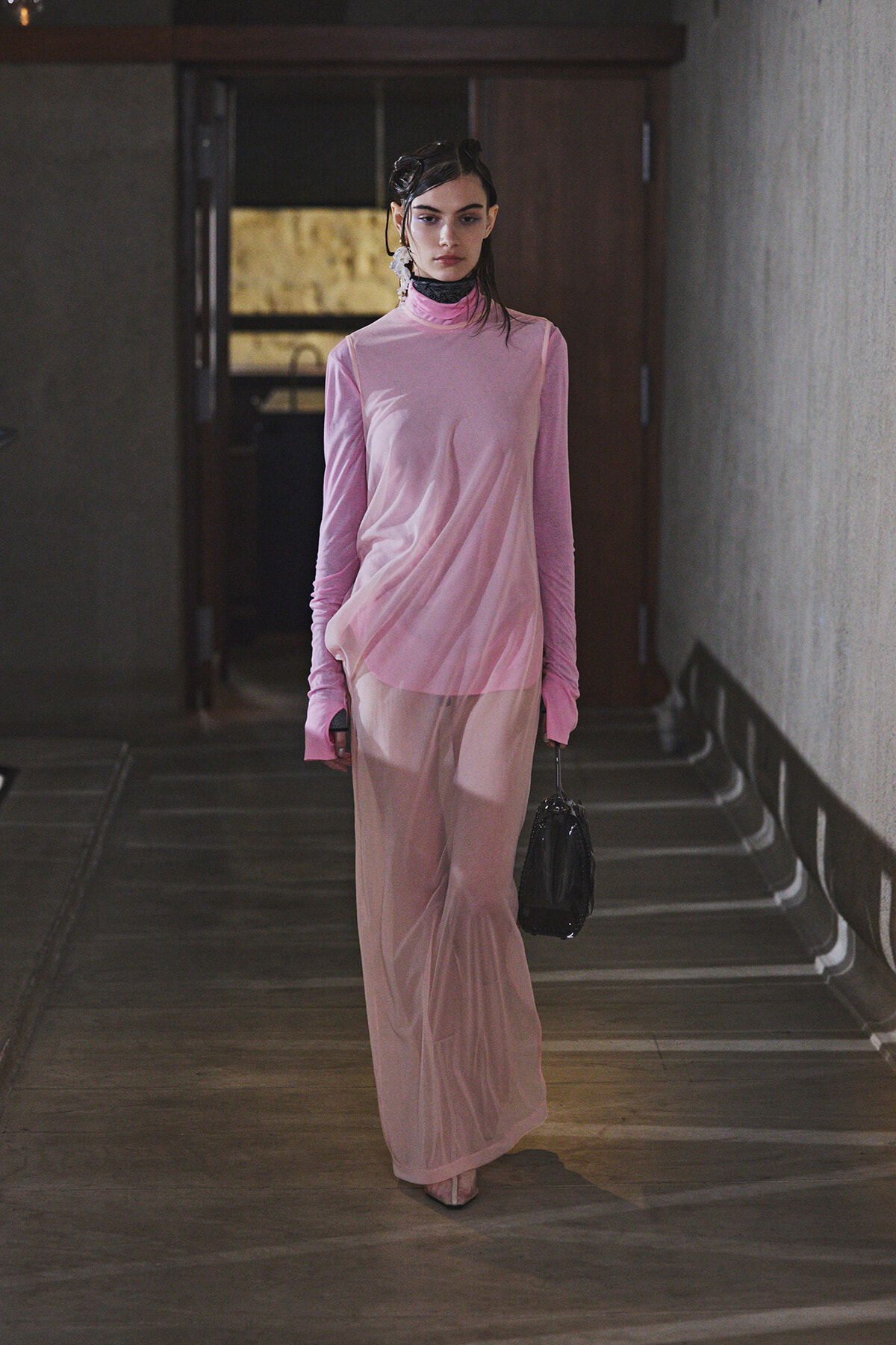 Model in a light pink, long-sleeve, high-neck dress walking down a dim hallway with a small black handbag.