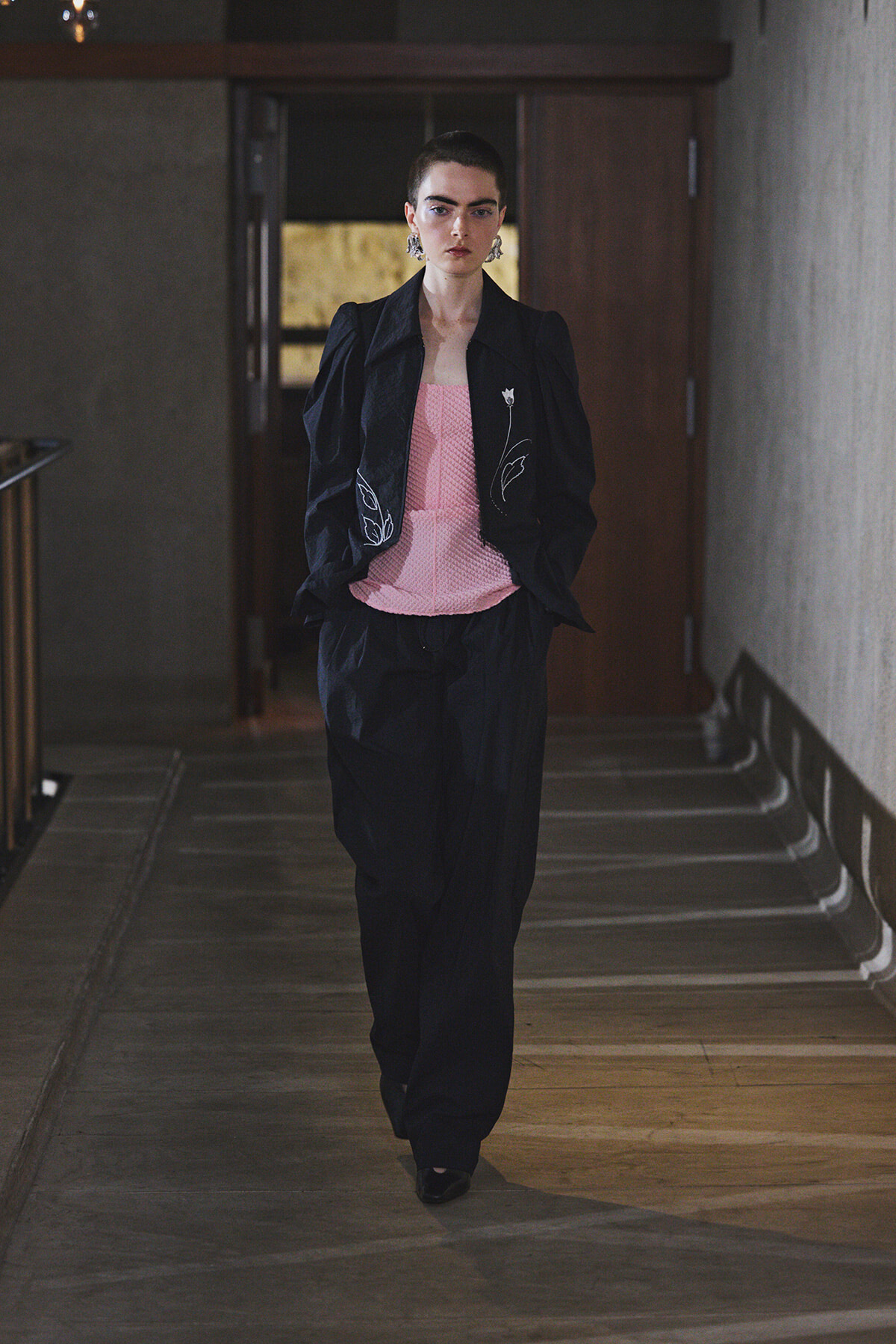 Model walking down a dim hallway in a black suit with a pink textured top and embroidered leaf details, wearing statement earrings.