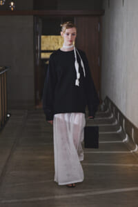 Model walking down a dim hallway in an oversized black sweater, a sheer white star-patterned skirt, a white scarf tied around the neck, and carrying a small black handbag.