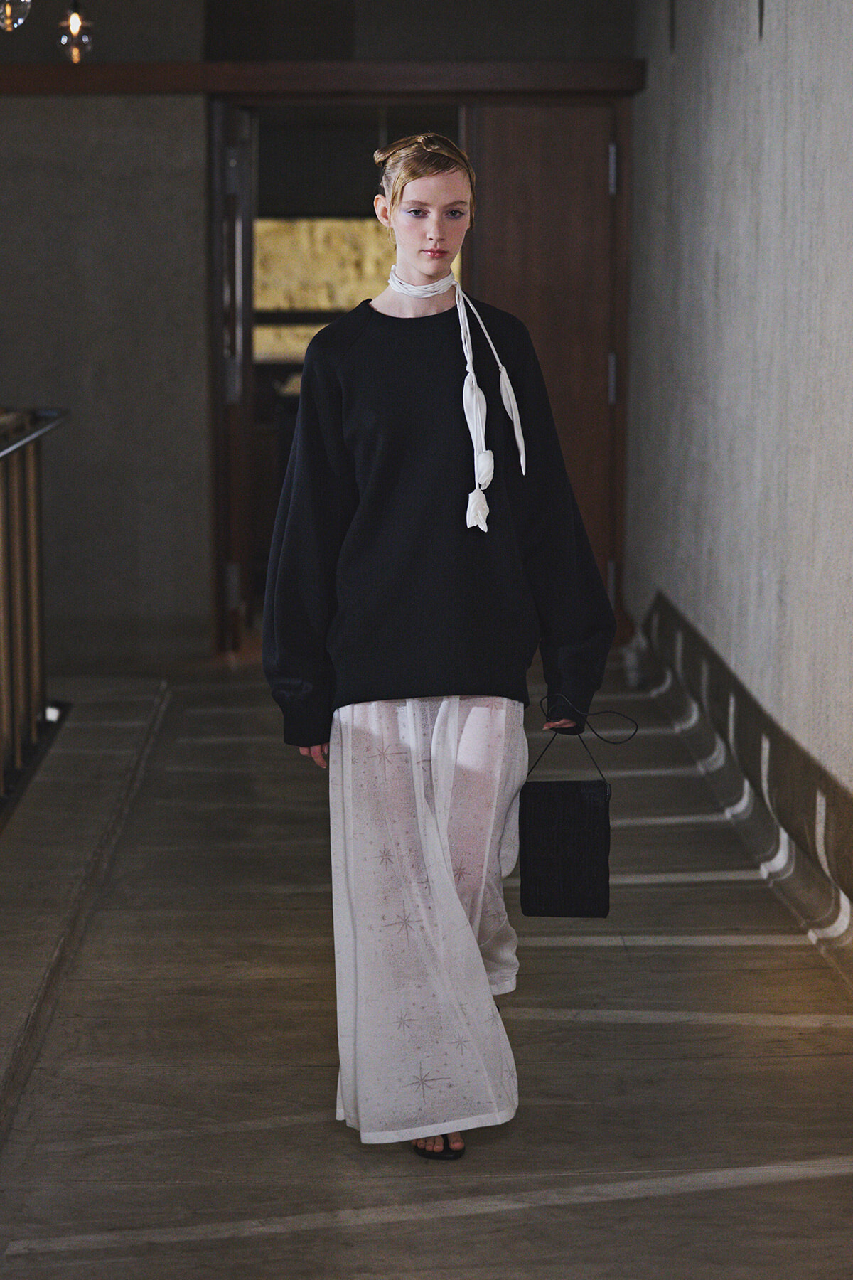 Model walking down a dim hallway in an oversized black sweater, a sheer white star-patterned skirt, a white scarf tied around the neck, and carrying a small black handbag.