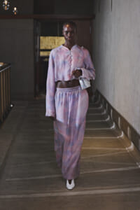 Model wearing pastel purple-pink tie-dye cropped blouse and matching wide-leg pants, carrying a white bag over the shoulder in a dim hallway.