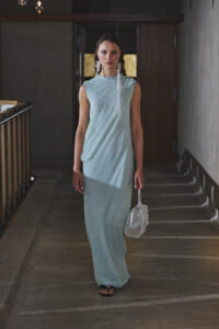 Woman in a pale blue sleeveless maxi dress walks down a dim hallway, holding a small white purse.