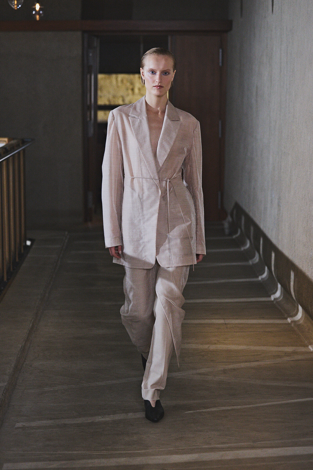 Model walking down a hallway in a light beige belted blazer and matching trousers, minimalist fashion look.