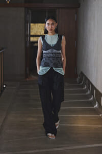 Model walking in a dim hallway wearing a layered dark patterned vest over a light blue top and black cargo pants.