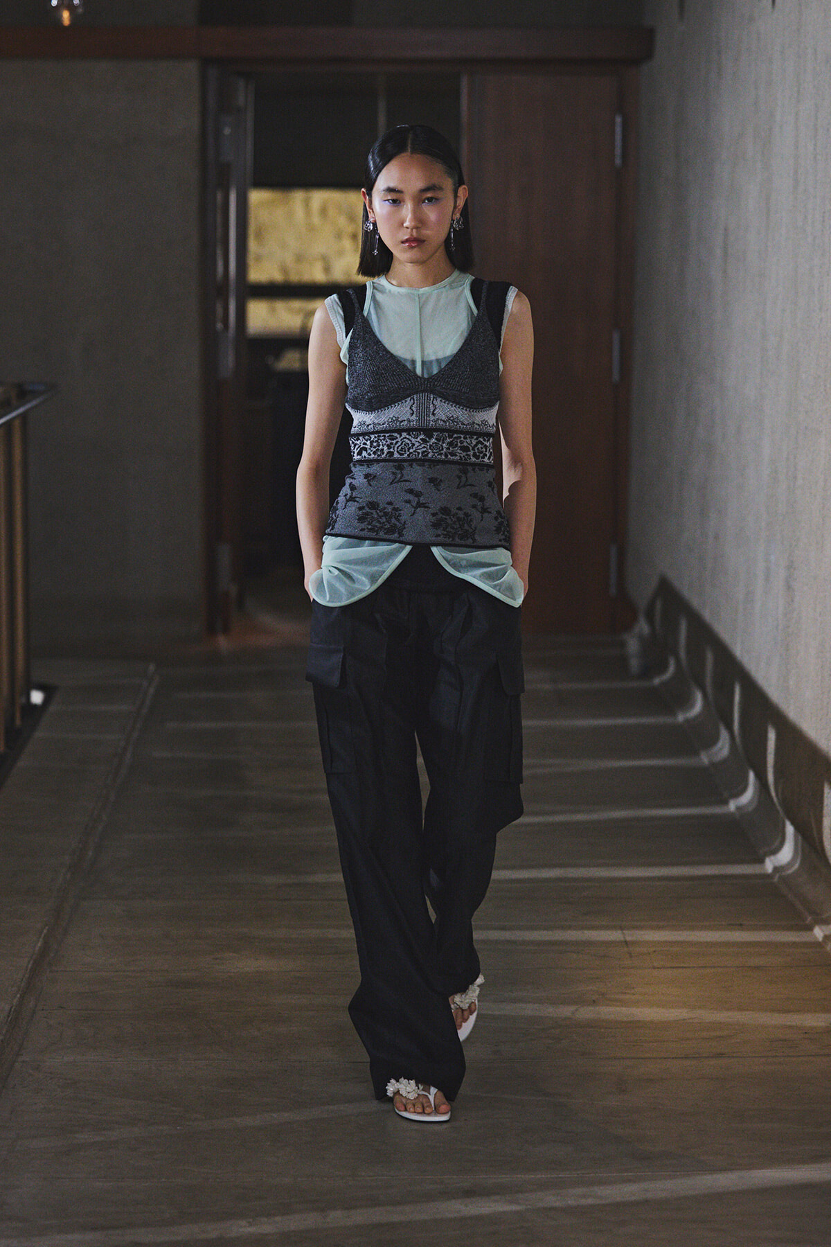 Model walking in a dim hallway wearing a layered dark patterned vest over a light blue top and black cargo pants.