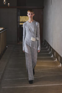 Model walking toward the camera in a silver lace long-sleeve dress, with a small pale crossbody bag, in a dim hallway runway setting.