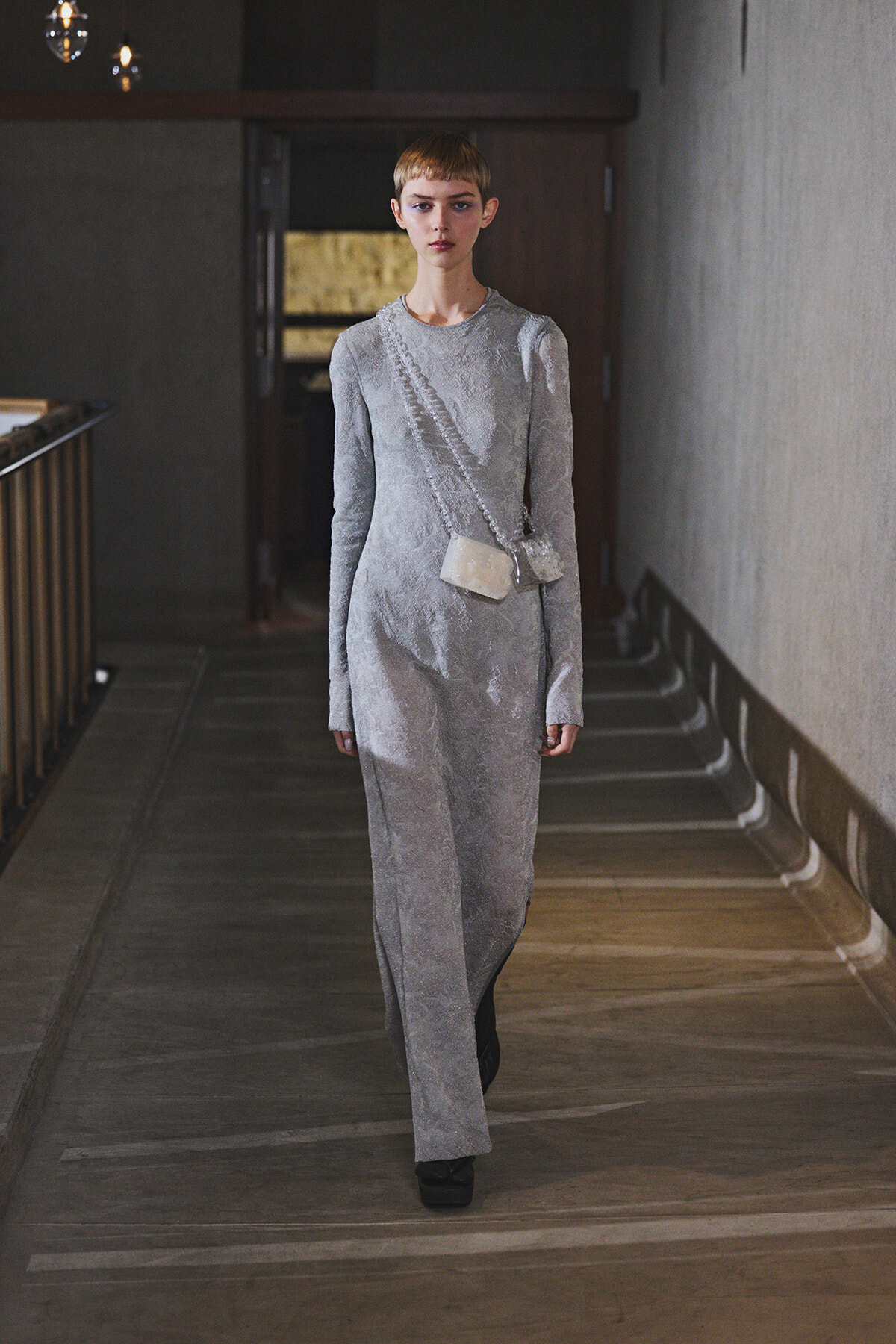 Model walking toward the camera in a silver lace long-sleeve dress, with a small pale crossbody bag, in a dim hallway runway setting.