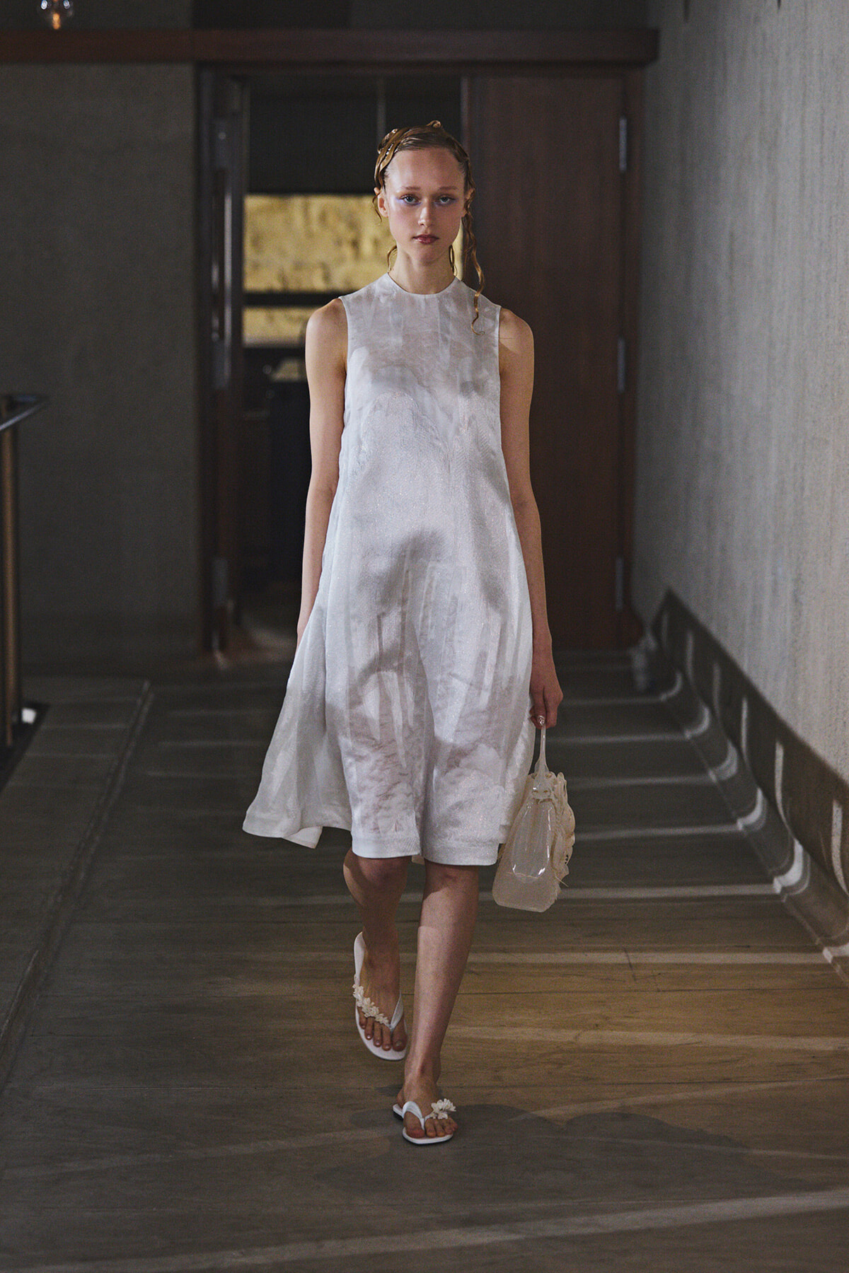 Model wearing a knee-length white sleeveless dress walks down a dim hallway, carrying a small white handbag and wearing white sandals.