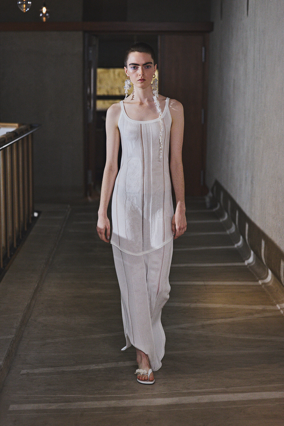 Model walking down a dim hallway in a white sleeveless dress with vertical panels, paired with wide-leg pants and large white earrings.