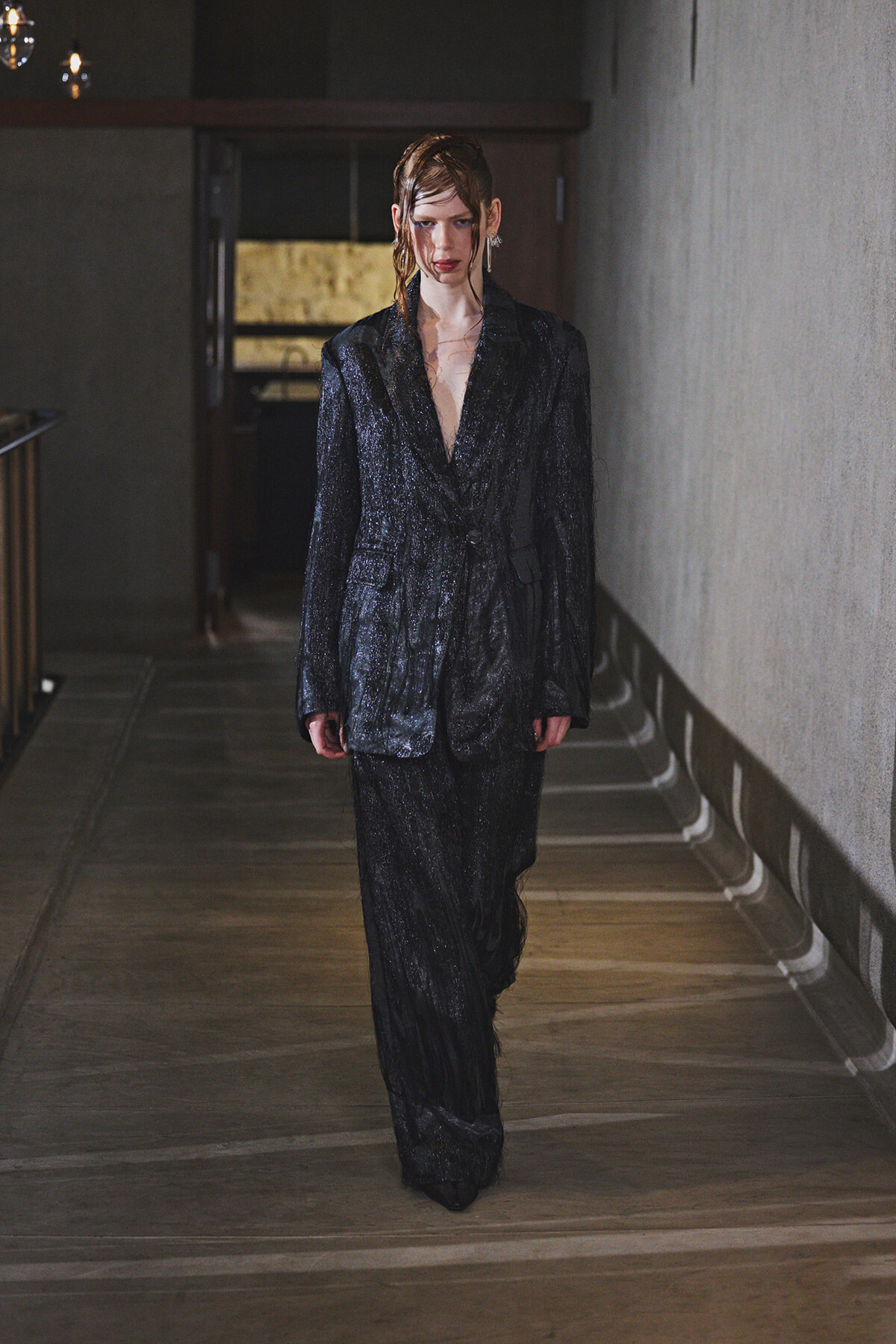 Model walking down a dim hallway wearing a dark, glittery blazer and matching pants; wet hair and bold makeup accent the look.
