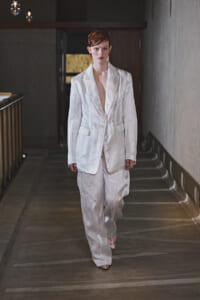 Model in a white suit walking down a dim hallway toward the camera, blazer open at the chest.