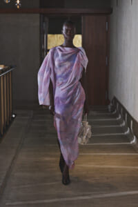 Full-length image of a dark-skinned model walking down a dim hallway in a pastel lavender-pink, draped gown, carrying a clear handbag.