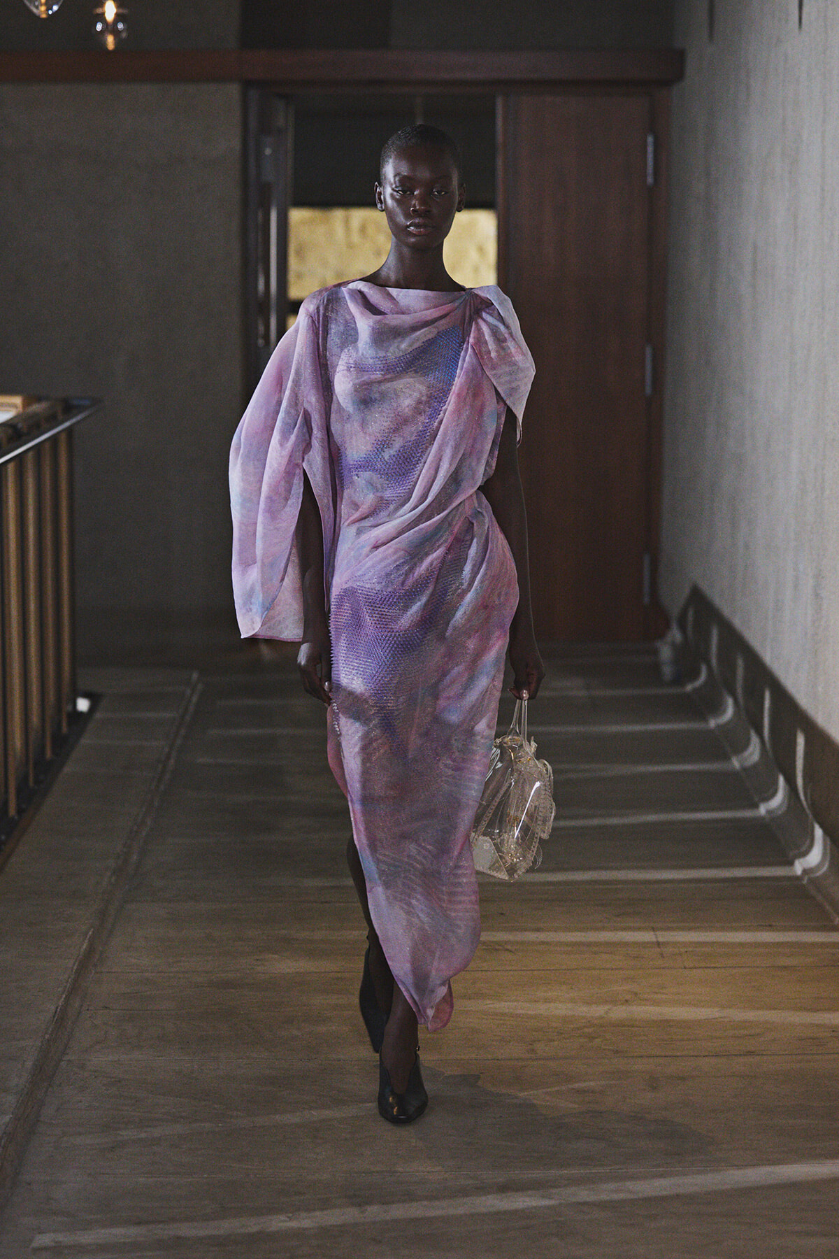 Full-length image of a dark-skinned model walking down a dim hallway in a pastel lavender-pink, draped gown, carrying a clear handbag.