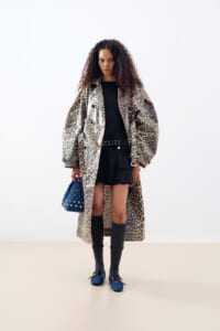 Model wearing a leopard-print long coat over a black top and skirt, with a studded belt, knee-high socks, blue loafers, and a blue studded handbag.