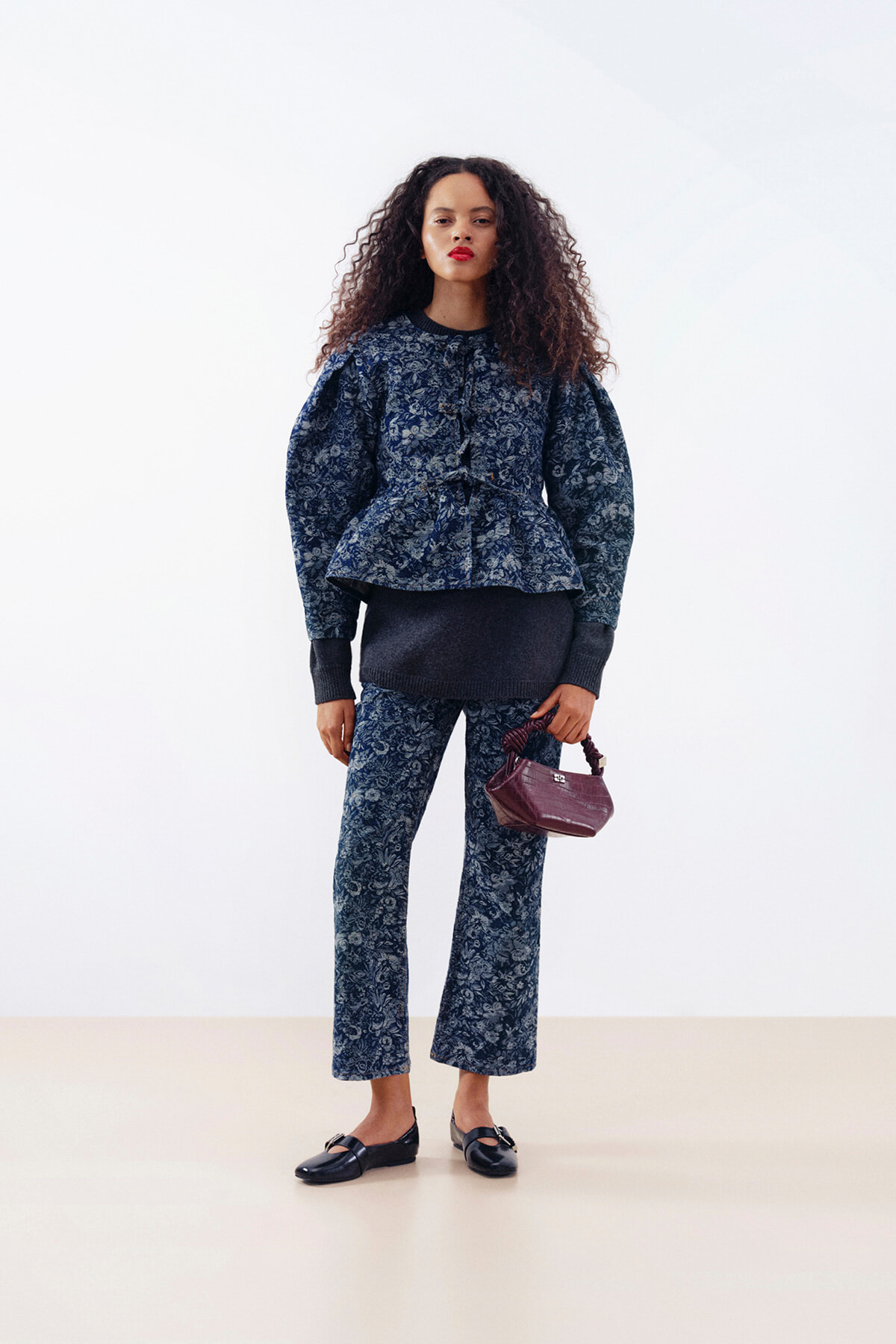 Woman with curly hair wearing a navy floral peplum top and matching wide-leg pants, black loafers, and a burgundy handbag.