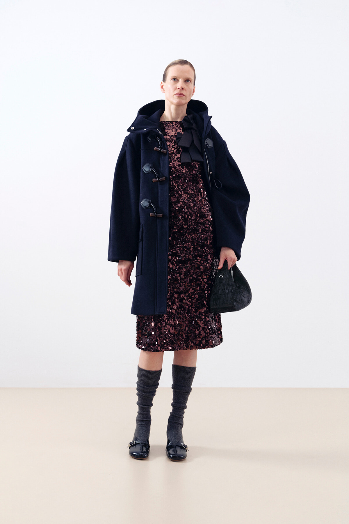 Model wearing a navy duffle coat over a burgundy sequin dress, with gray leg warmers, black loafers, and a black handbag in a studio setting.