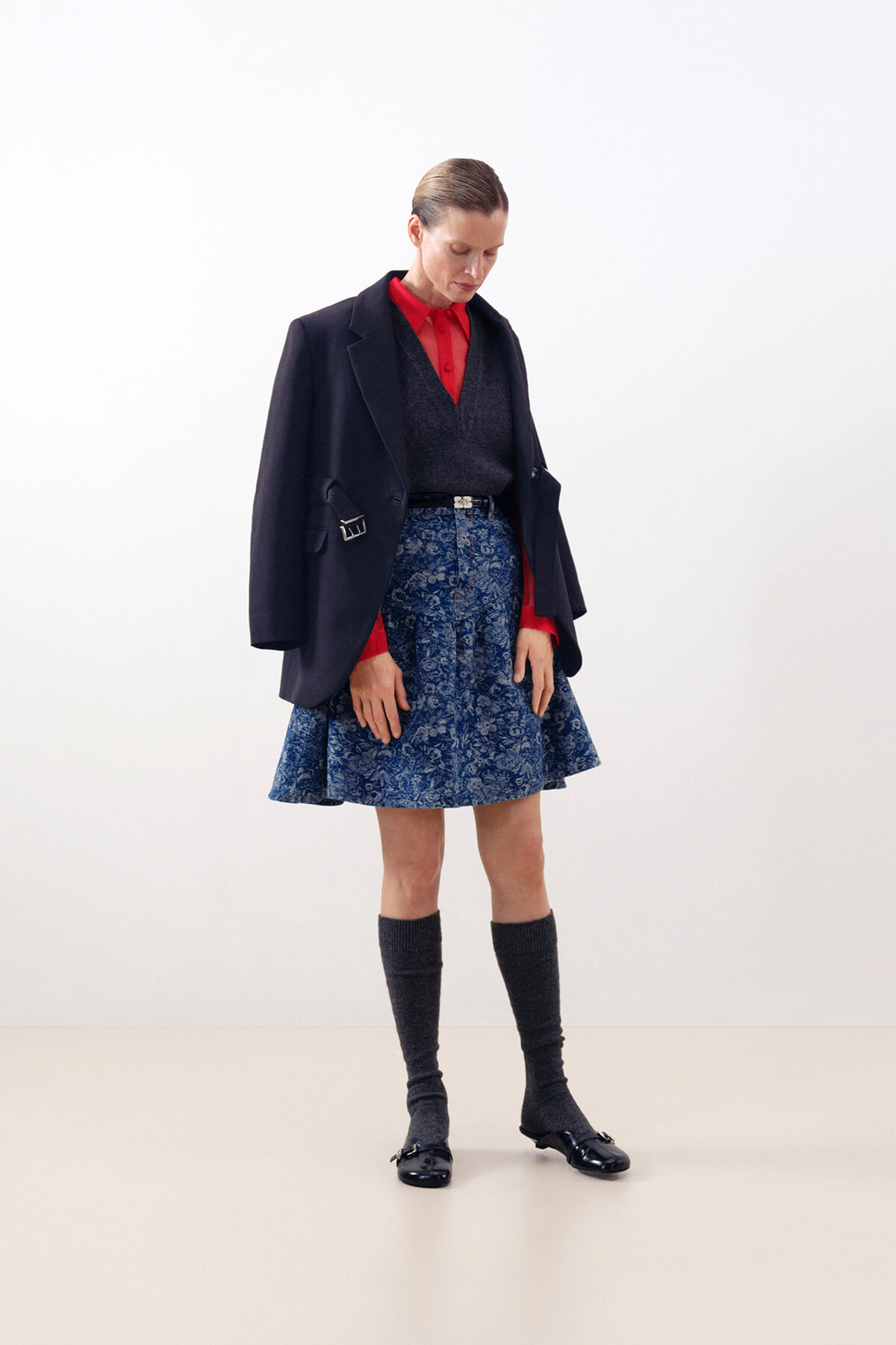 Model wearing a navy blazer over a red shirt with a blue floral A-line skirt, knee-high socks, and black flats against a light backdrop.