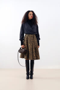 Woman in a dark wrap sweater over a black top, leopard-print midi skirt with a wide belt, black knee-high boots, and a small brown handbag.