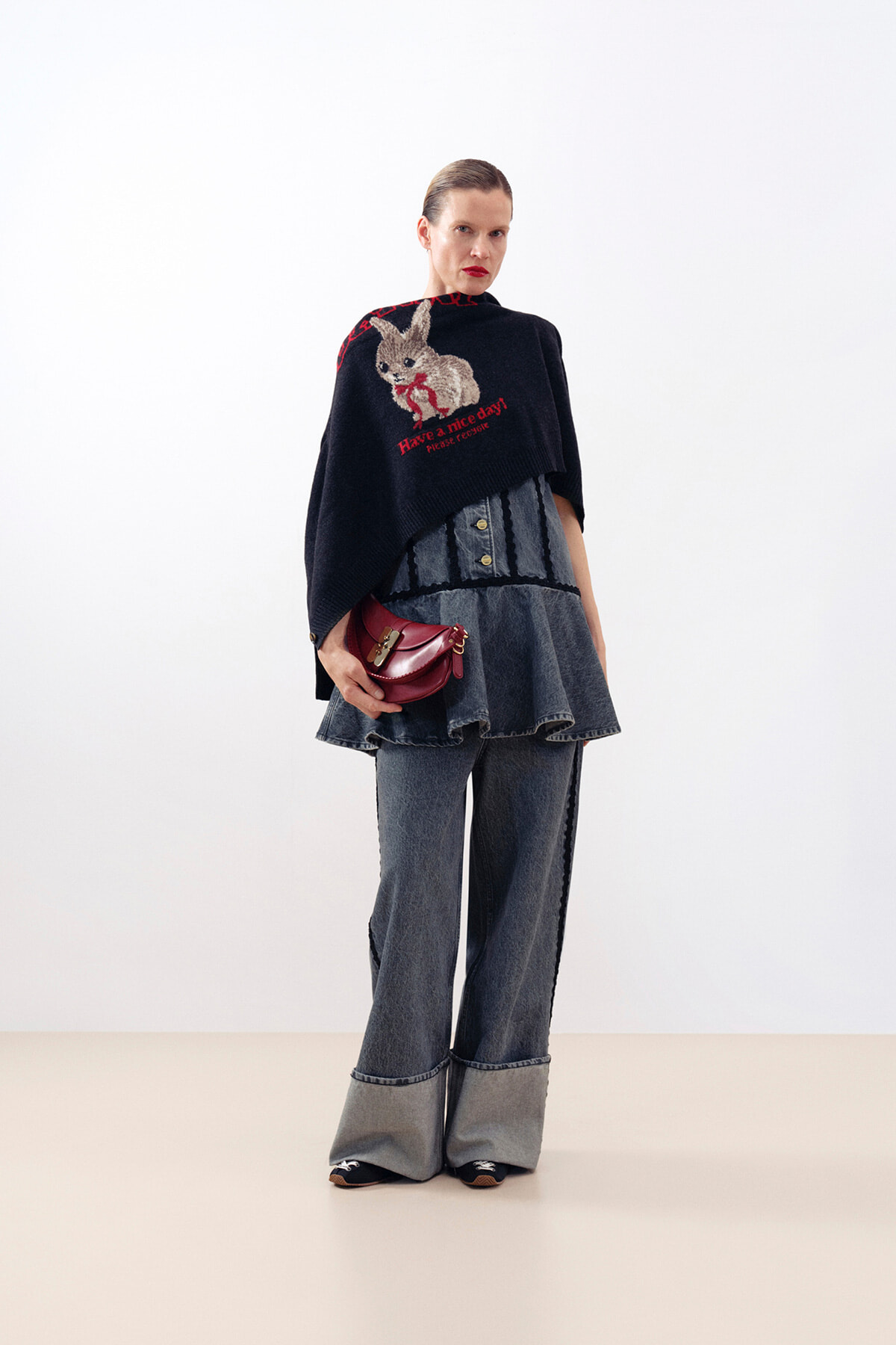Model wearing a black poncho with a rabbit graphic over a layered denim top, wide-leg jeans, and a burgundy handbag in a studio setting.