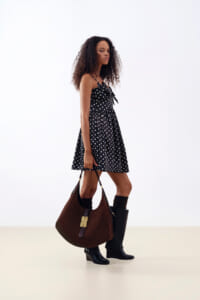 Side view of a woman in a black polka-dot dress with a bow detail, carrying a large brown handbag and wearing black knee-high boots.