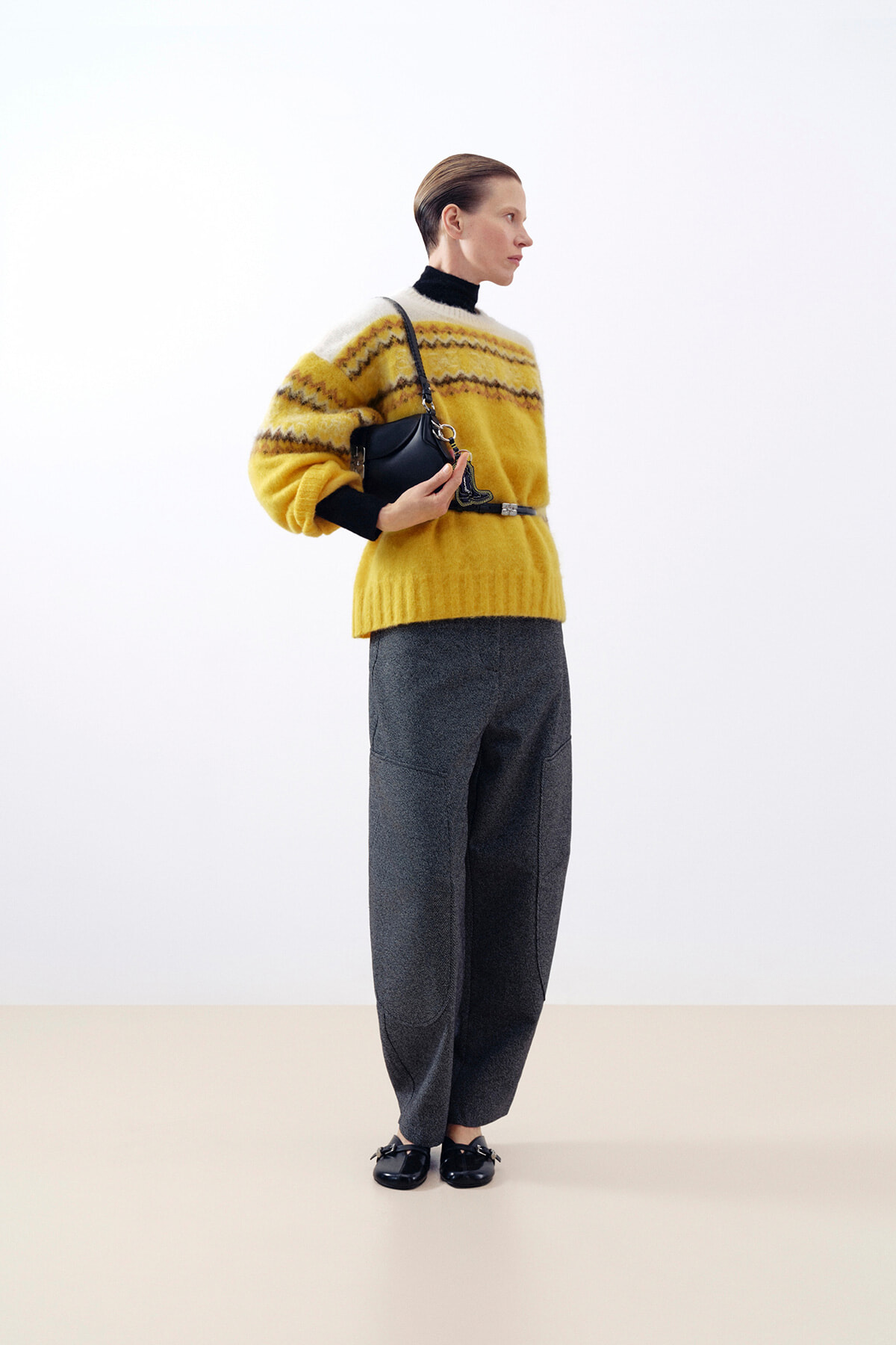 Model wearing a bright yellow knit sweater with a patterned yoke, paired with gray wide-leg pants and a small black shoulder bag in a studio setting