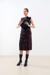Model wearing a burgundy sequin midi dress with a large black bow at the shoulder, holding a dark clutch against a light backdrop.