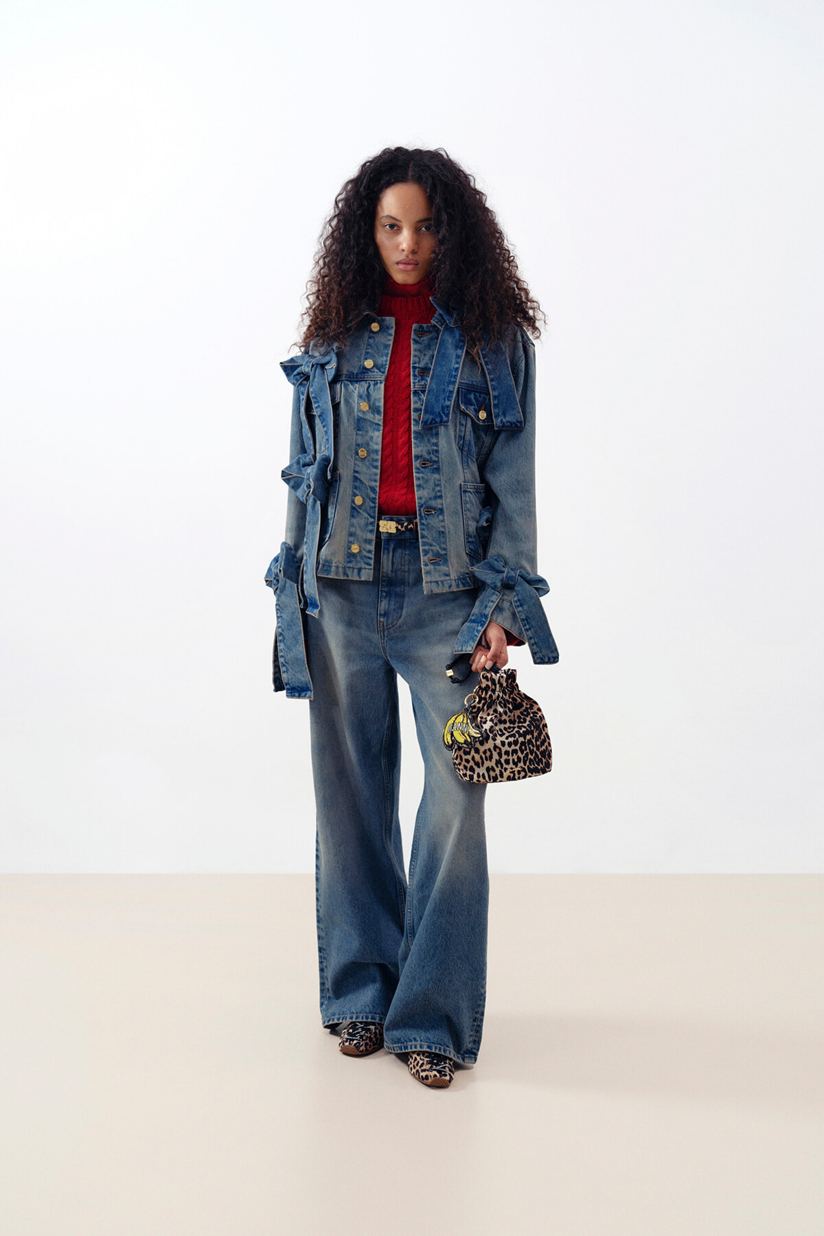 Model in a denim-on-denim outfit: blue jacket and wide-leg jeans, red knit turtleneck, and leopard-print handbag and shoes, standing against a pale backdrop.