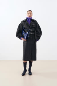 Person wearing a long dark coat with a purple turtleneck, holding a blue handbag at the waist.