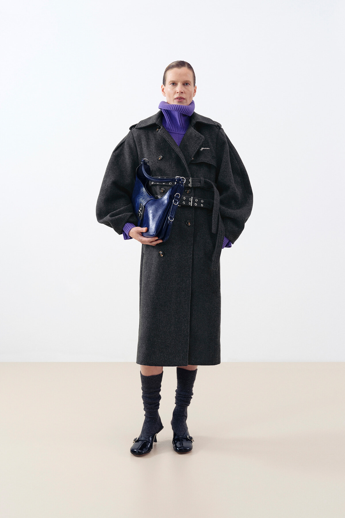 Person wearing a long dark coat with a purple turtleneck, holding a blue handbag at the waist.