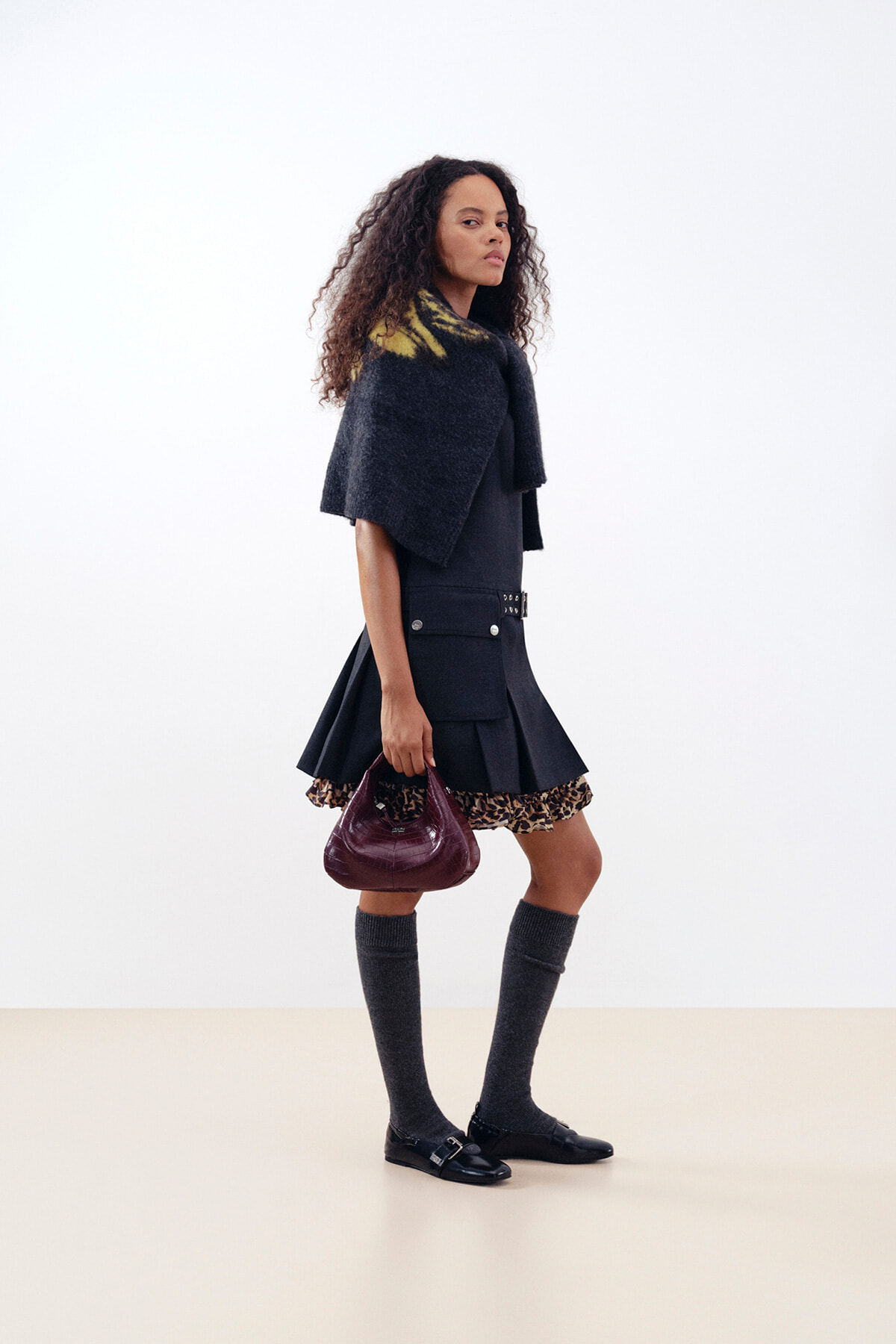 Side view of a fashion model walking, wearing a dark capelet over a black outfit with a pleated skirt that has a leopard-print hem, knee-high gray socks, black loafers, and a burgundy handbag.