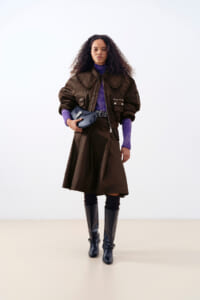 Woman with curly hair wearing a brown oversized coat, purple top, and black knee-high boots, holding a blue clutch while standing against a light background.
